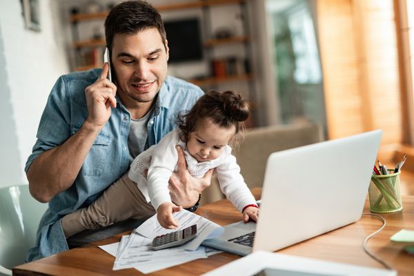 Young working father talking on the phone while babysitting his playful daughter at home.