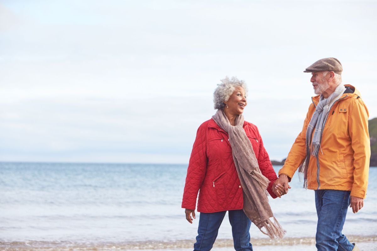 Mixed race senior couple walking on beach holding hands, smiling at one another