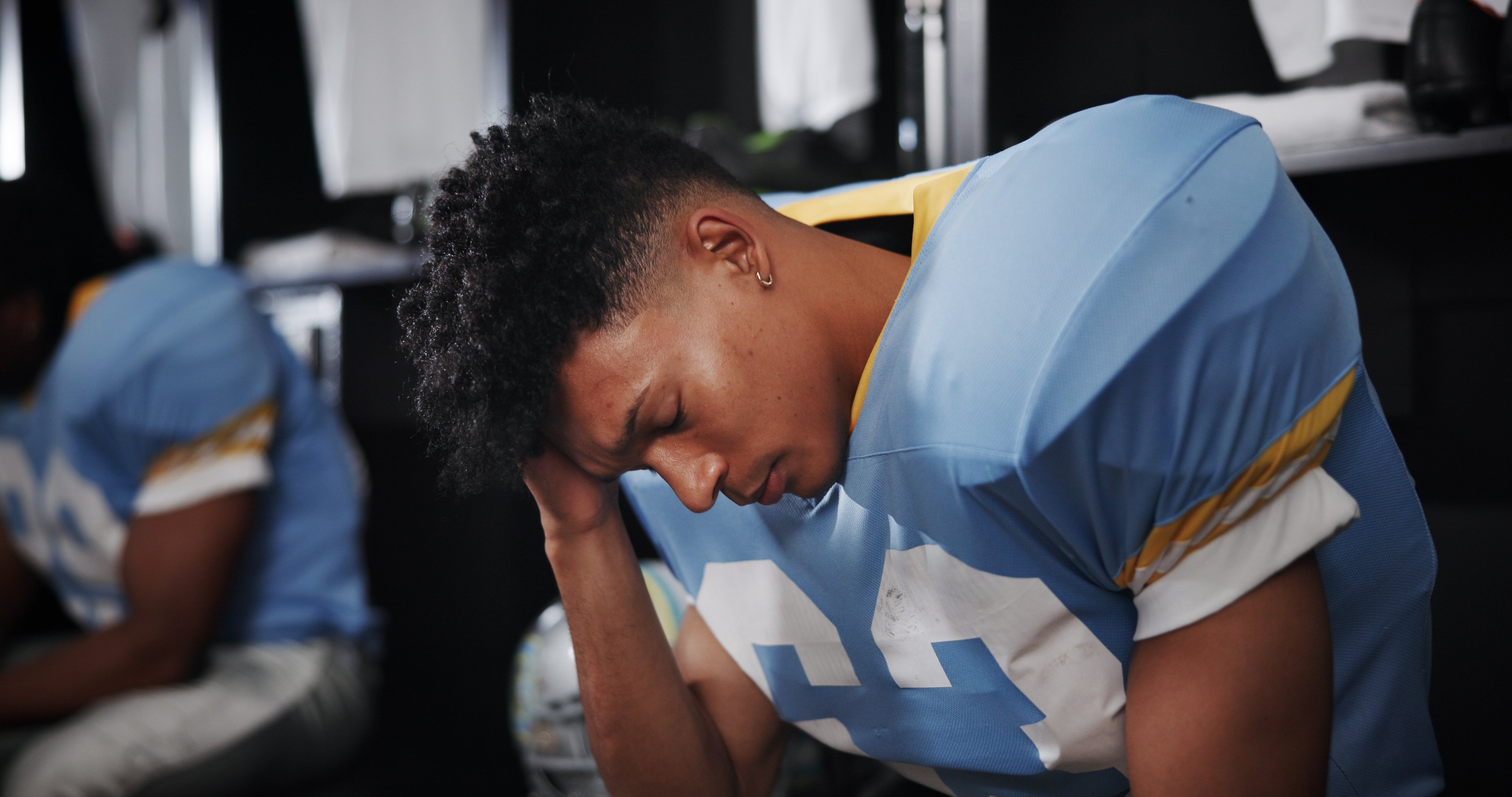 young black male football athlete in locker room holding his head in his hands